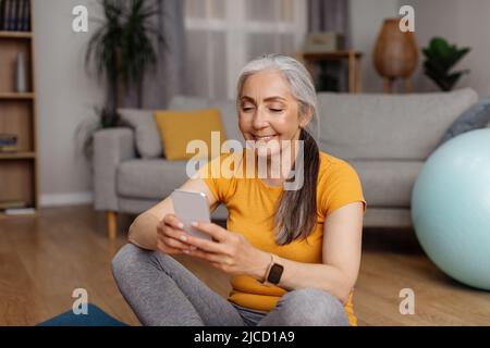 Donna anziana in forma felice che si rilassa sul tappetino yoga con smartphone dopo il suo allenamento a casa, prendendo pausa dopo la pratica yoga Foto Stock