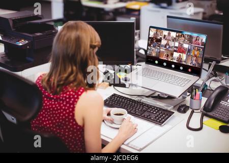 Donna d'affari caucasica che tiene una tazza di caffè video conferenza con i colleghi sul laptop in ufficio Foto Stock