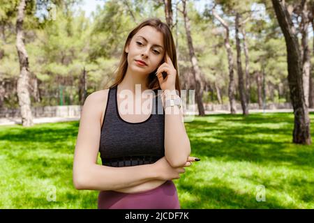 Giovane donna bruna che indossa abiti sportivi nel parco cittadino, all'aperto tocca la testa con le dita in profondità nei pensieri, pensando a cose importanti. Speranza. Sadnes Foto Stock