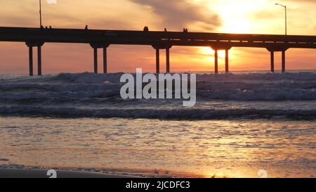 Silhouette di persone che camminano, molo su pali in acqua di mare. Onde oceaniche, cielo drammatico al tramonto. California costa estetica, spiaggia o spiaggia vibrazione al tramonto. Estate stagcape a San Diego vicino Los Angeles Foto Stock