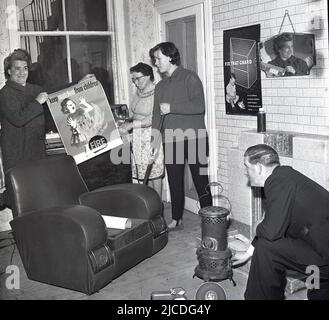 1960s, storica, campagna di educazione alla sicurezza antincendio, all'interno di una stanza, una donna che tiene un fuoco elettrico e un uomo inginocchiato da una piccola stufa, in una foto per evidenziare la prevenzione del fuoco in casa. Una donna tiene un poster che avverte del pericolo delle partite..'tenere le partite lontano dai bambini', mentre un altro poster è sul muro che sottolinea l'importanza delle guardie antincendio...' fissa quella guardia (fuoco)', Fife, Scozia, Regno Unito. Foto Stock