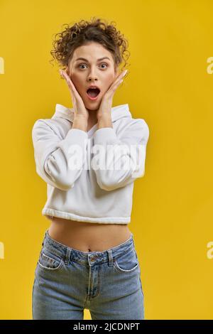 Bella donna in felpa con cappuccio, coprendo il viso con le mani, guardando la macchina fotografica con la bocca agape al chiuso. Ritratto vista di ragazza carina, scioccato o spaventata, isolato su sfondo arancione studio. Concetto di emozioni. Foto Stock