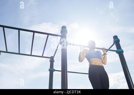 Determinata ragazza in abbigliamento sportivo stretto facendo pull-up sul bar strutture alla luce del sole mentre si pratica l'allenamento all'aperto Foto Stock