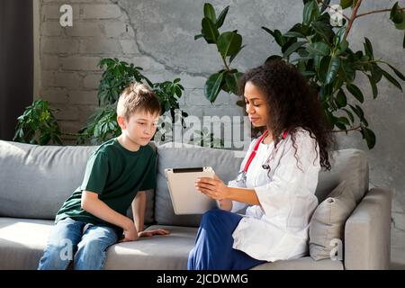 Pediatra donna con pelle scura che usa l'applicazione medica per lavorare con i pazienti pediatrici e ottenere il ragazzo di storia medica in ufficio medico. Controllo regolare. Medi Foto Stock