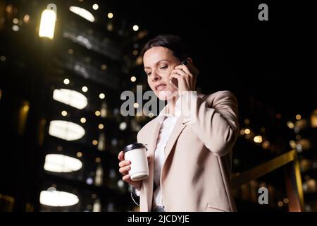 Sorridente donna d'affari di mezza età in giacca che tiene una tazza di caffè e parla al telefono mentre si sposta giù le scale della hall di notte Foto Stock