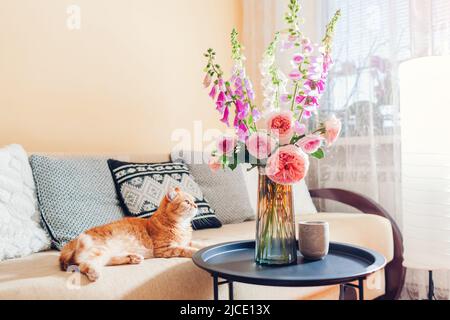 Gatto zenzero sdraiato sul divano in soggiorno da bouquet di rose fresche e fiori di foxguanto. Gli animali domestici sono comodi e accoglienti Foto Stock