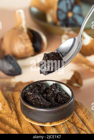 Cucchiaio con pasta d'aglio nera. Una piccola ciotola di argilla con aioli neri su un piatto di legno fatto a mano. Bulbo fermentato all'aglio sullo sfondo. Sano Foto Stock