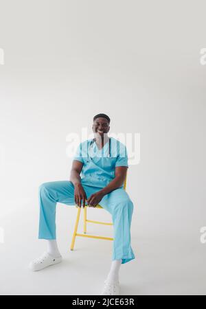 Un medico africano in una tuta medica turchese si siede su una sedia su un isolato sfondo bianco Foto Stock