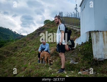 Cisneros, Antioquia, Colombia - 20 febbraio 2022: L'uomo bianco guarda e gode il paesaggio accanto ad un uomo sudamericano seduto e Petting un cane Foto Stock