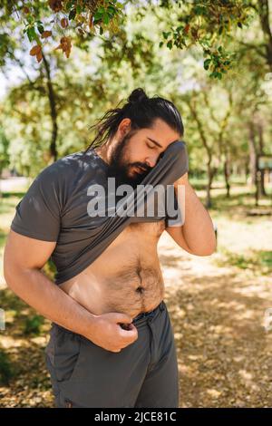 giovane uomo che pratica sport all'aperto pulendo il sudore dalla fronte con la sua t-shirt. atleta che si prepara per una maratona. salute e benessere stile di vita. Foto Stock