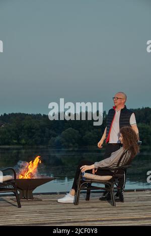 La coppia carina guarda il tramonto seduto accanto al fuoco sul molo Foto Stock