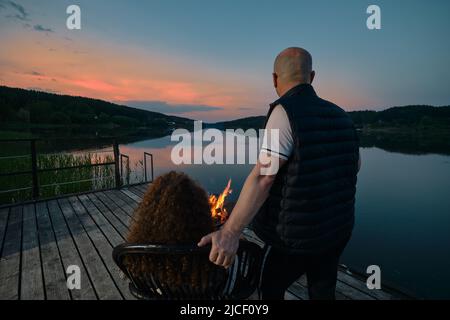 Simpatica coppia guarda il tramonto seduto accanto al fuoco sul molo (vista da dietro) Foto Stock