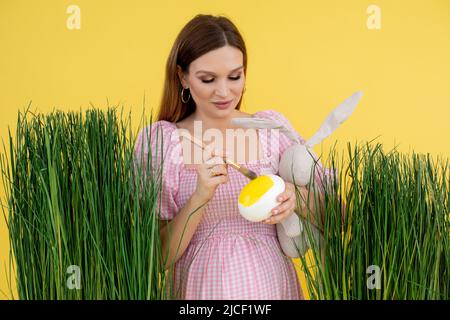 Donna felice in tarda gravidanza su decorazione erba vernice uovo con spazzola e tenere giocattolo morbido coniglio, sfondo giallo. Ritratto di giovane donna incinta Foto Stock