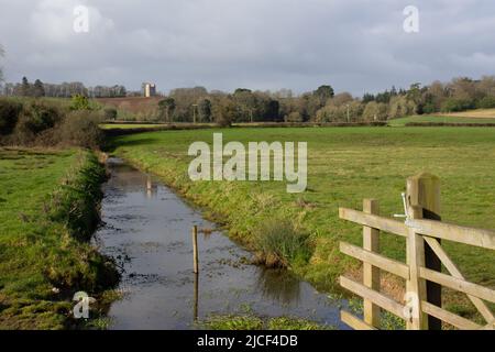 Canale di drenaggio e cancello in tipico terreno agricolo inglese con Folly in lontananza e un cielo blu e nuvole bianche Foto Stock