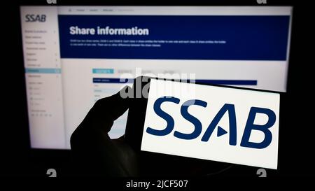 Persona che tiene il telefono cellulare con il logo della società siderurgica svedese SSAB AB sullo schermo di fronte alla pagina web aziendale. Mettere a fuoco sul display del telefono. Foto Stock