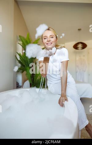 Allegra giovane donna bionda caucasica seduta in vasca da bagno e giocando con schiuma e bolle. Foto Stock
