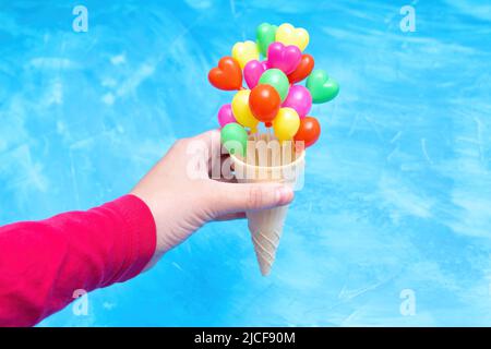 Mano che tiene un cono di cialda gelato con palloncini a forma di cuore a forma di festa contro uno sfondo blu con spazio copia. Foto Stock