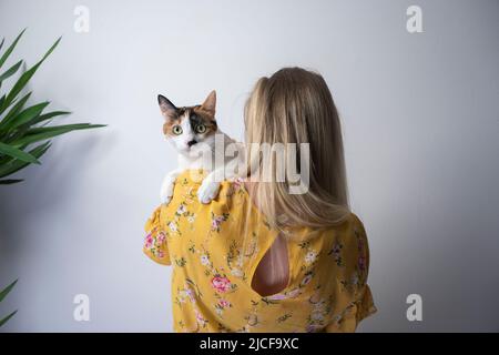 vista posteriore della giovane donna bionda vestito giallo con motivo floreale che porta carino gatto calico a spalla Foto Stock