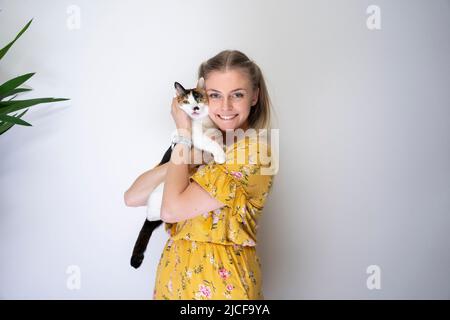 giovane donna bionda che porta il gatto bianco calico sulle braccia abbracciando il kitty sorridendo alla macchina fotografica guardando felice con lo spazio di copia Foto Stock