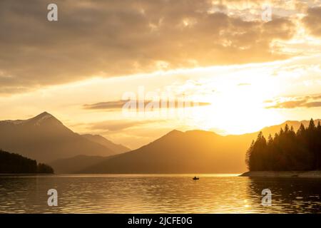 Atmosfera serale poco prima del tramonto a Walchensee nelle Alpi bavaresi, in primo piano un pescatore in una barca a remi, sullo sfondo il Simetsberg, l'atmosfera nuvolosa e il tramonto del sole. Foto Stock