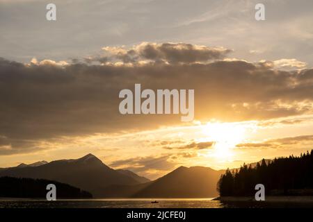 Atmosfera serale poco prima del tramonto a Walchensee nelle Alpi bavaresi, in primo piano un pescatore in una barca a remi, sullo sfondo il Simetsberg, l'atmosfera nuvolosa e il tramonto del sole. Foto Stock