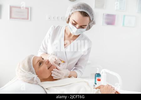 Cosmetologo grazioso sta facendo l'iniezione alla donna caucasica bella anziana in gabinetto Foto Stock