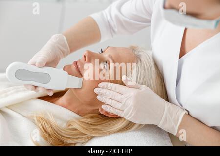 Il cosmetologo professionale sta facendo la pulizia ultrasonica del viso per bella donna matura caucasica felice in clinica Foto Stock