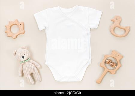 Bodysuit bianco avvolgente per mocappo bambino. Mockup di vestiti per bambini su sfondo marrone pastello con giocattoli e accessori per bambini. Battiti e teet di legno Foto Stock