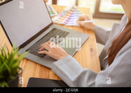 Giovane grafica femminile asiatica che lavora nel suo ufficio, utilizzando un computer portatile per controllare il suo progetto. Immagine ritagliata Foto Stock