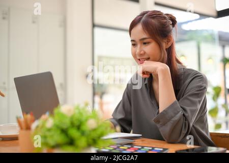 Creativa giovane designer grafico asiatico femminile o interior designer utilizzando un computer portatile, lavorando alla sua scrivania in ufficio in camera d'ufficio Foto Stock