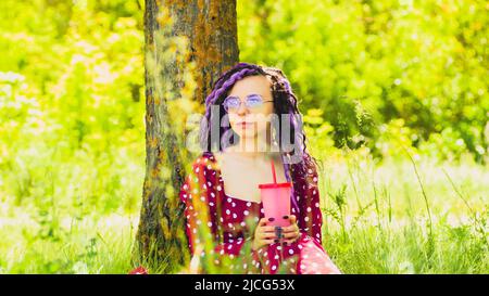 Giovane donna in occhiali, rosso vestito bere bevanda rinfrescante, seduta vicino albero in soleggiato, vento, tempo estivo. Bella femmina bere soft drink t Foto Stock