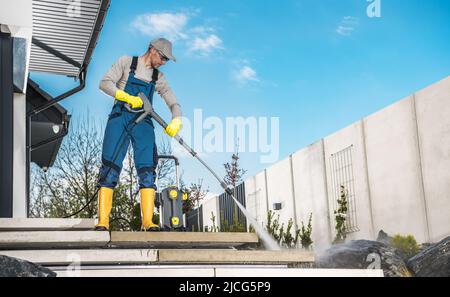 Uomini caucasici nel suo 40s lavatrice il suo giardino cortile sentieri in cemento e scale utilizzando la idropulitrice professionale. Foto Stock