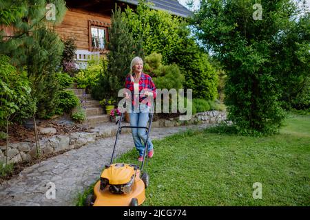 Donna anziana che taglia erba con tosaerba in giardino, concetto di lavoro in giardino. Foto Stock