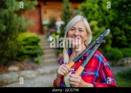 Ritratto di donna anziana giardinaggio in estate, tenendo potare sheares e guardando la macchina fotografica. Foto Stock