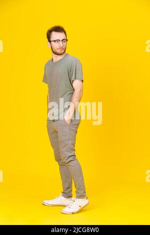 Il giovane uomo indossa occhiali da vista casual in posa su sfondo giallo con le mani in tasche che guardano lateralmente. Elegante uomo hipster dal look casual elegante. Ragazzo premuroso su sfondo giallo. Foto Stock