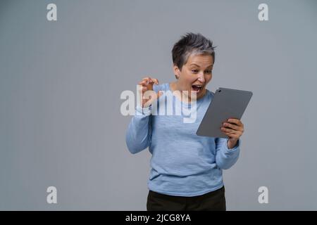 Donna d'affari ispirata e scioccata con capelli grigi maturi e tablet digitale in mano al lavoro, allo shopping, al gioco online. Bella donna nel 50s in blusa blu isolato su bianco. Foto Stock