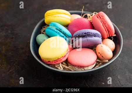 Macaroon multicolore e uova di cioccolato in ciotola di ceramica su sfondo di cemento nero. Vista laterale, primo piano, natura morta. Colazione, mattina, concetto. Foto Stock