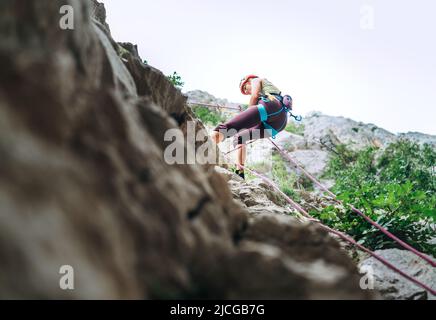 Donna arrampicatrice attiva in casco protettivo che si abbella dalla parete rocciosa della scogliera utilizzando la corda con dispositivo belay e imbracatura da arrampicata. Tempo attivo per gli sport estremi Foto Stock