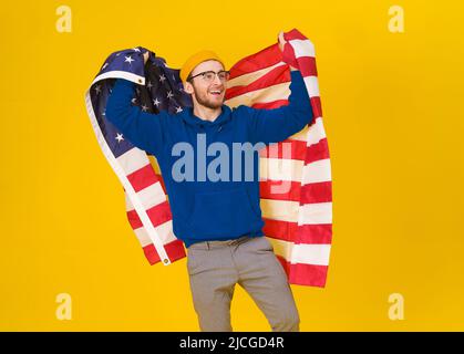 Il bel giovane caucasico celebra il 4th luglio in felpa blu con cappuccio e bandiera degli Stati Uniti dietro la schiena isolato su sfondo giallo. La libertà è nella vostra vita. Giovane allegro con bandiera americana. Foto Stock