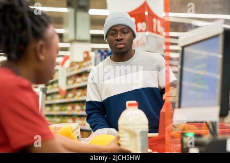 Giovane consumatore maschio serio in casualwear in piedi dal banco di cassa in supermercato, mentre venditore scansione prodotti alimentari Foto Stock