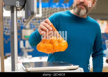 Uomo maturo con barbetta grigia da putting con mandarini freschi su bilance elettroniche mentre li pesa nel reparto alimentari Foto Stock