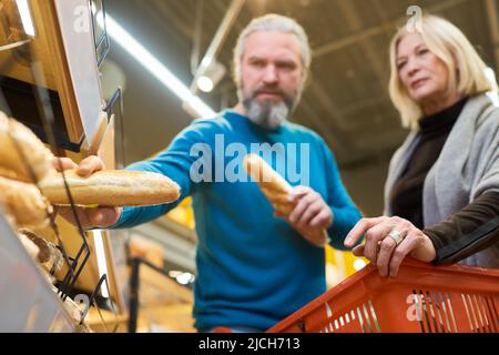 Cliente maschio maturo che prende due baguette di grano fresco dallo scaffale in supermercato mentre si trova accanto a sua moglie con carrello Foto Stock