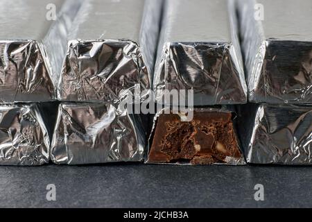 Tavolette di cioccolato fondente con noci tritate, avvolte in un foglio accatastato su un tavolo scuro. Primo piano. Giorno di cioccolato fondente Foto Stock