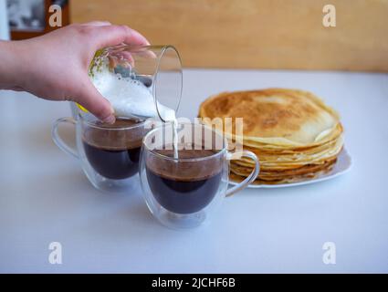 Chiudere versando il latte da un bicchiere in una tazza con caffè appena fatto con frittelle su un piatto di sfondo Foto Stock