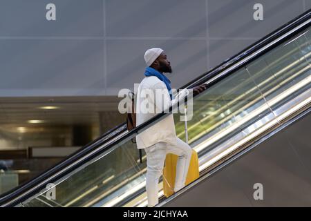 Passeggero afro-americano con valigia stand sulla scala mobile, tiene corrimano nel terminal dell'aeroporto Foto Stock