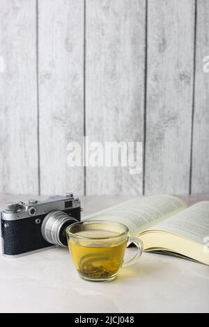 Tè verde alle erbe in una tazza trasparente accanto a un libro aperto e una macchina fotografica vintage su sfondo chiaro Foto Stock