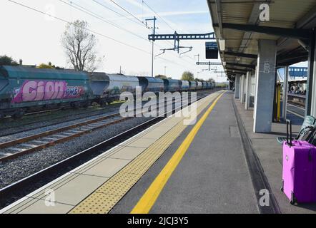 Piattaforma ferroviaria britannica con binari vuoti e valigie in attesa di un viaggio con un treno cargo sullo sfondo. Foto Stock