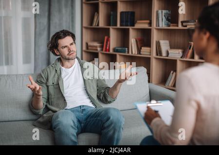 Triste insoddisfatto uomo europeo stende le braccia ai lati e ascolta la psicologa nera femminile Foto Stock