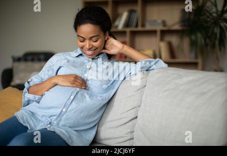 Happy incinta African American Woman sorridente stroking Belly seduta al coperto Foto Stock