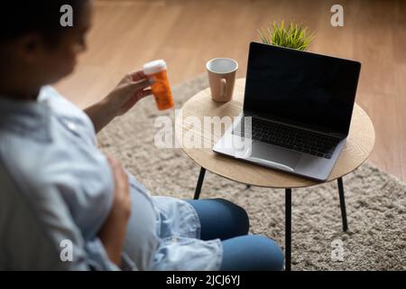 Donna nera incinta usando le pillole di tenuta del laptop a casa, corto Foto Stock